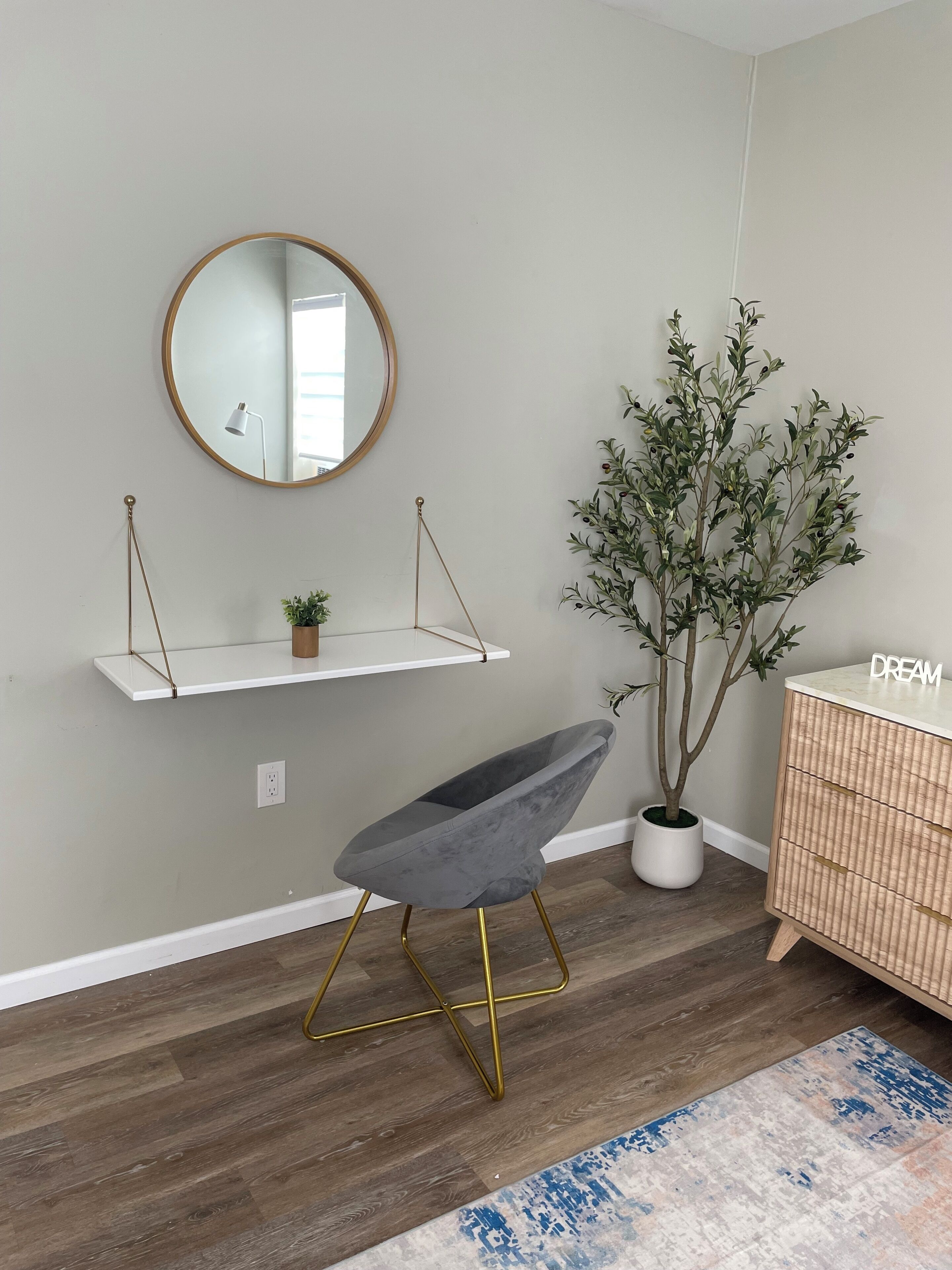 A modern room with a grey upholstered chair, a wooden floor, a white shelf with a small plant, a large mirror, and a wooden dresser with the word "DREAM" on it.