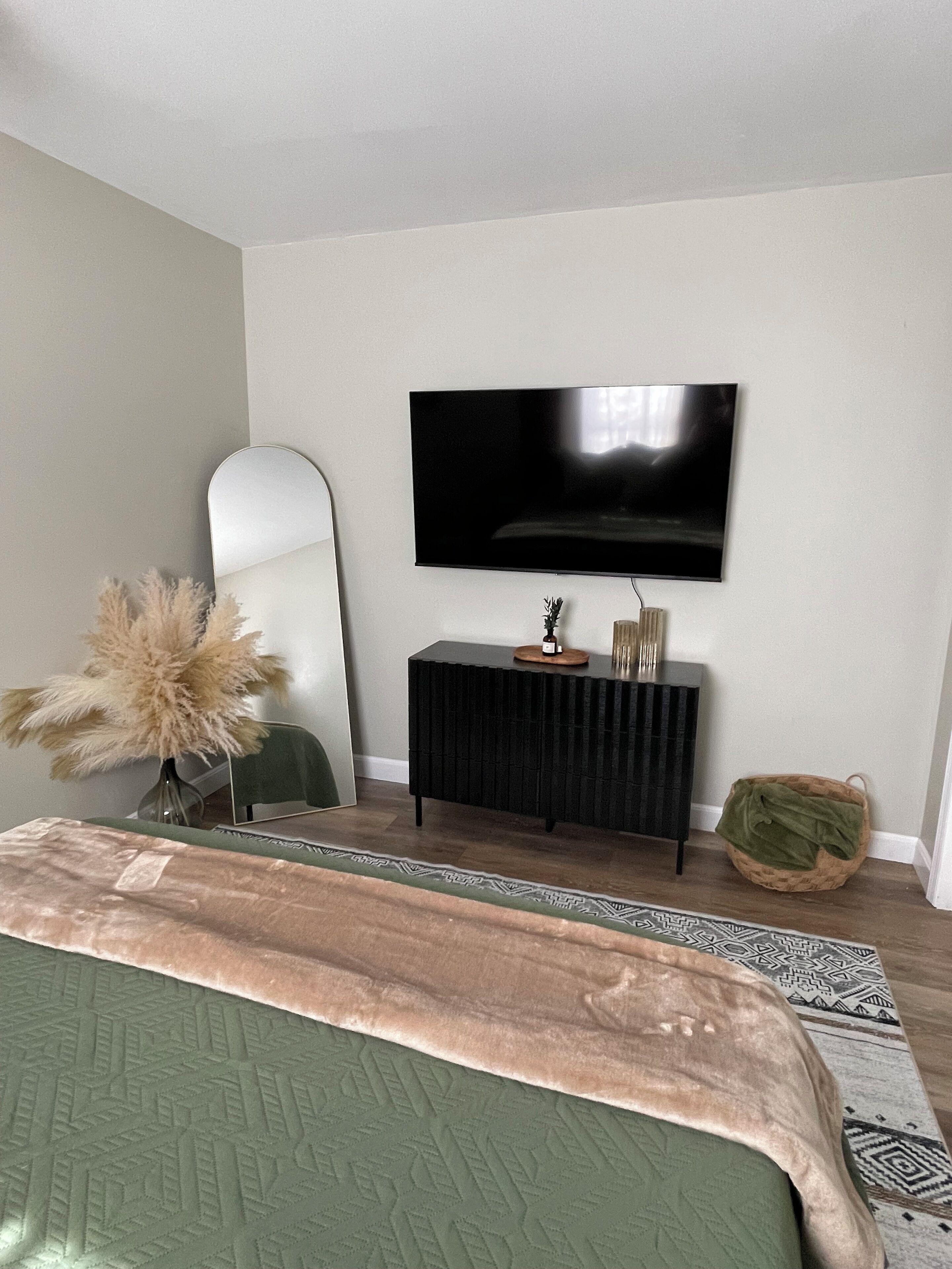 A bedroom with a bed, a TV mounted on the wall, a side table with decorative items, a mirror, and a patterned rug.