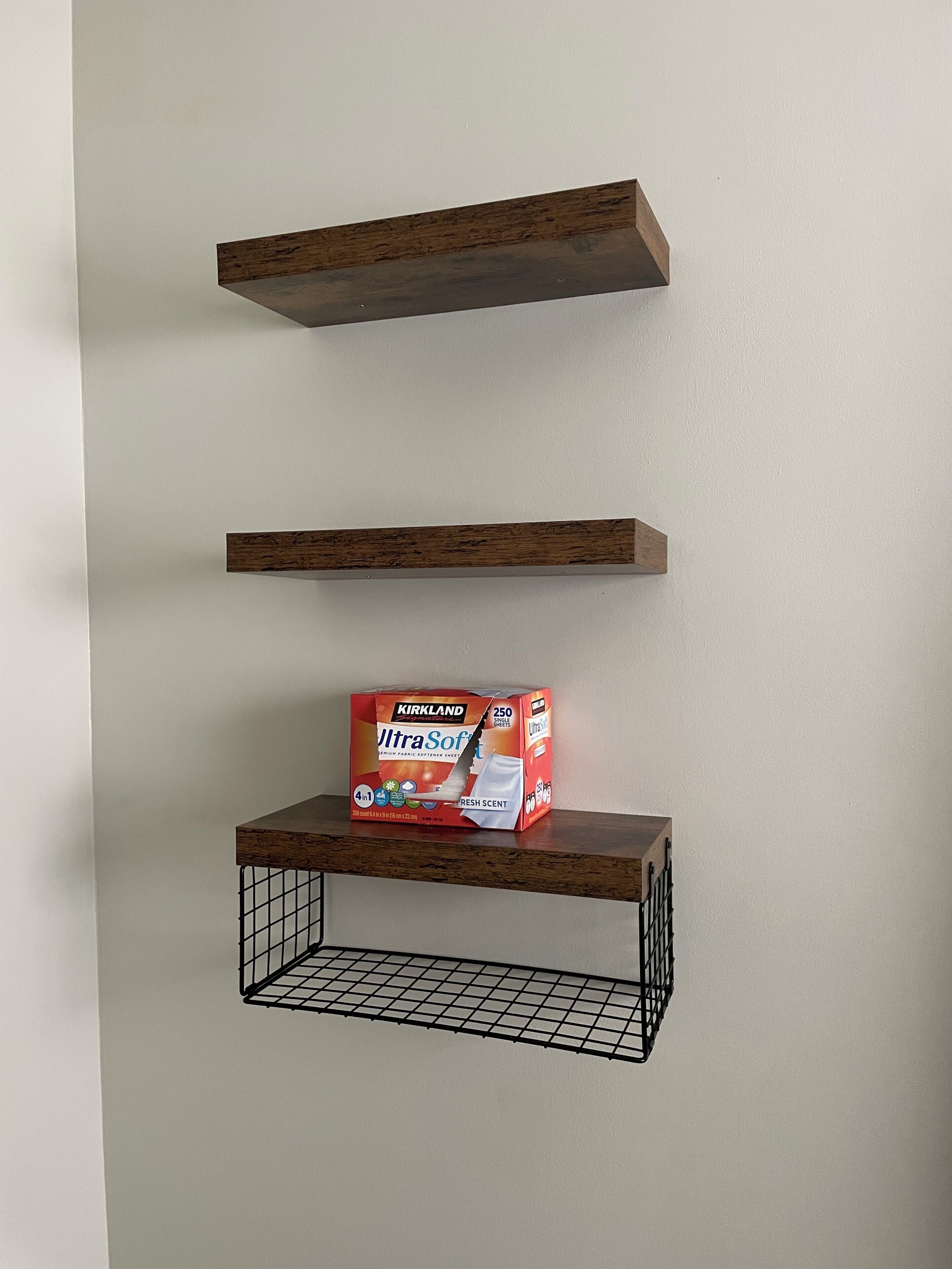 Uma parede com três prateleiras de madeira, uma delas com uma embalagem de amaciante de tecidos Kirkland UltraSoft.