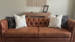 A brown leather sofa with tufted backrest and decorative pillows, two wooden side tables with bowls, a macramé wall hanging, and a framed welcome sign.