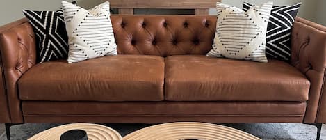 A brown leather sofa with tufted backrest and decorative pillows, two wooden side tables with bowls, a macramé wall hanging, and a framed welcome sign.
