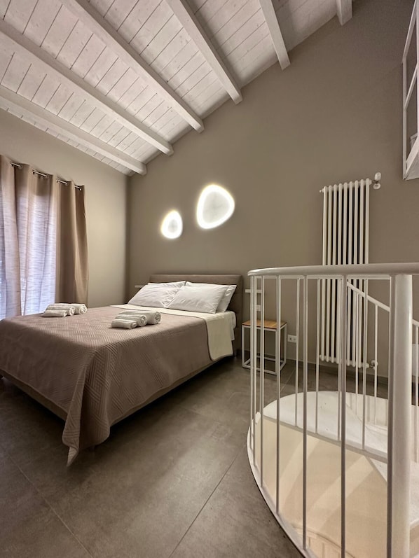 A bedroom with a bed, a white railing, a radiator, and a ceiling with wooden beams.