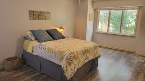 Un dormitorio con cama, mesita de noche, lámpara, ventana y una obra de arte colgada en la pared.