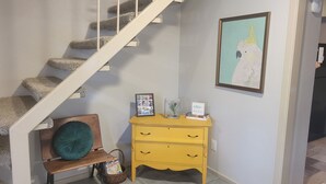 Una escalera con peldaños alfombrados, un banco de madera con cojín verde, un aparador amarillo con cajones, un cuadro enmarcado de un pájaro y una cesta con libros.