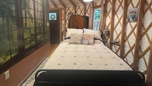 Une chambre avec un grand lit, des poutres en bois, une fenêtre donnant sur la forêt et une petite table avec une lampe.