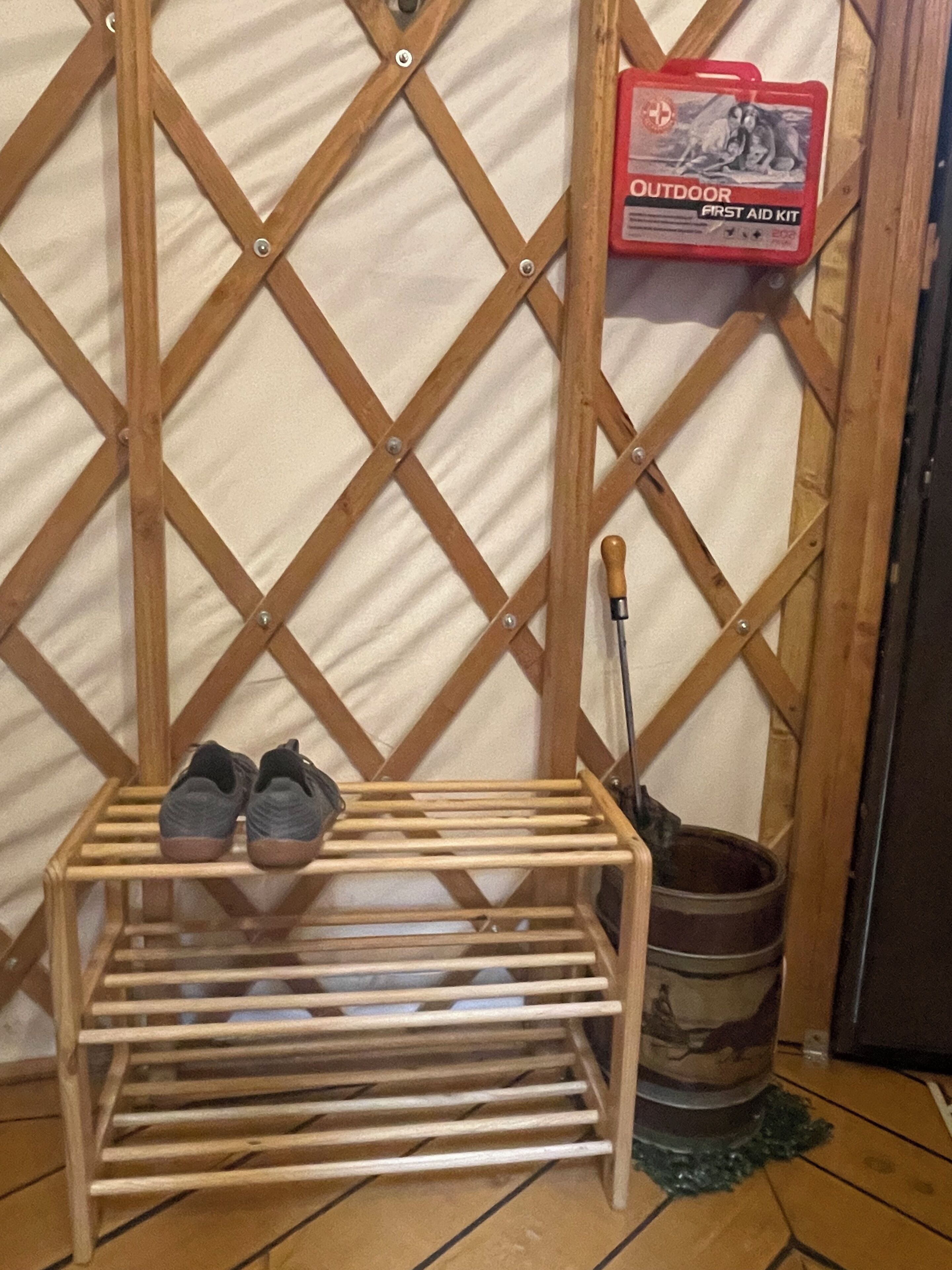Un porte-chaussures en bois avec une paire de chaussures, une trousse de premiers secours rouge pour l’extérieur et une serpillière appuyée contre le mur.
