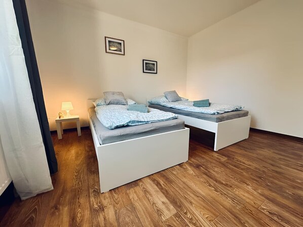 A room with two white beds, a small bedside table with a lamp, and a wooden floor.