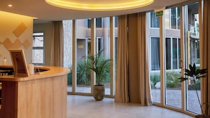 Eine Hotelrezeption mit Holztresen, einer großen Pflanze in einem Topf und Blick auf den Innenhof durch große Fenster.
