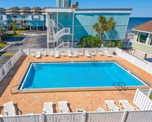 Ein rechteckiger Swimmingpool mit weißen Liegestühlen darum herum, eine Treppe, die zu einem Gebäude mit einem 'E' darauf führt, und Blick auf den Ozean.