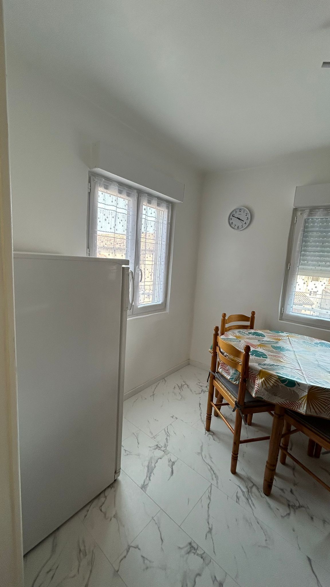 A compact kitchen with a refrigerator, a dining table with chairs, and two windows with blinds.