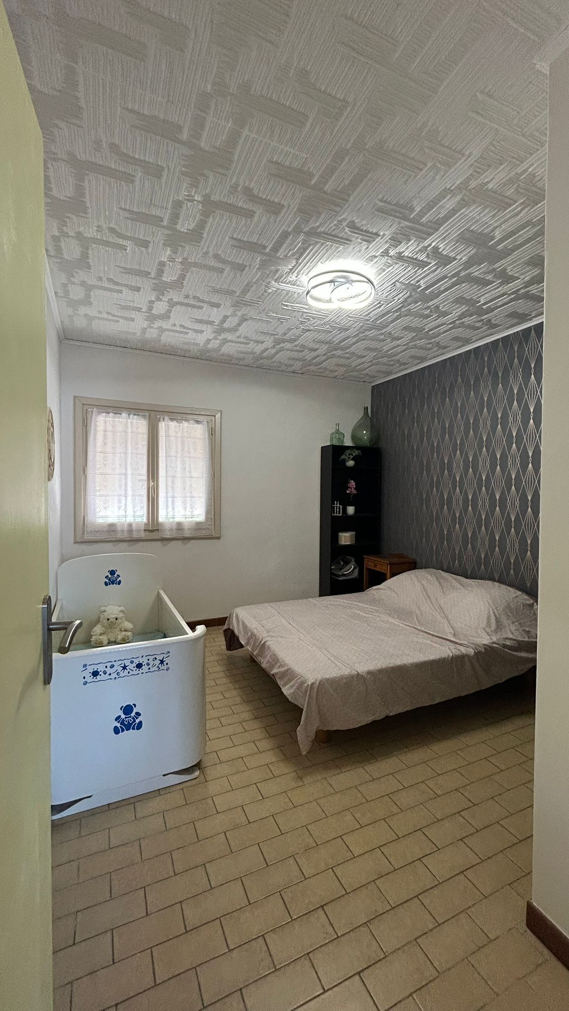 A bedroom with a single bed, a white bathtub with blue designs, a window with curtains, and a patterned wall.