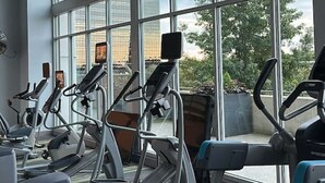 Een sportschool met loopbanden, hometrainers en elliptische trainers, grote ramen en uitzicht op gebouwen.