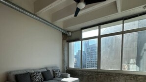 Une pièce dotée d’un ventilateur de plafond, d’une grande fenêtre donnant sur des immeubles, d’un canapé gris et d’un plancher en bois foncé.