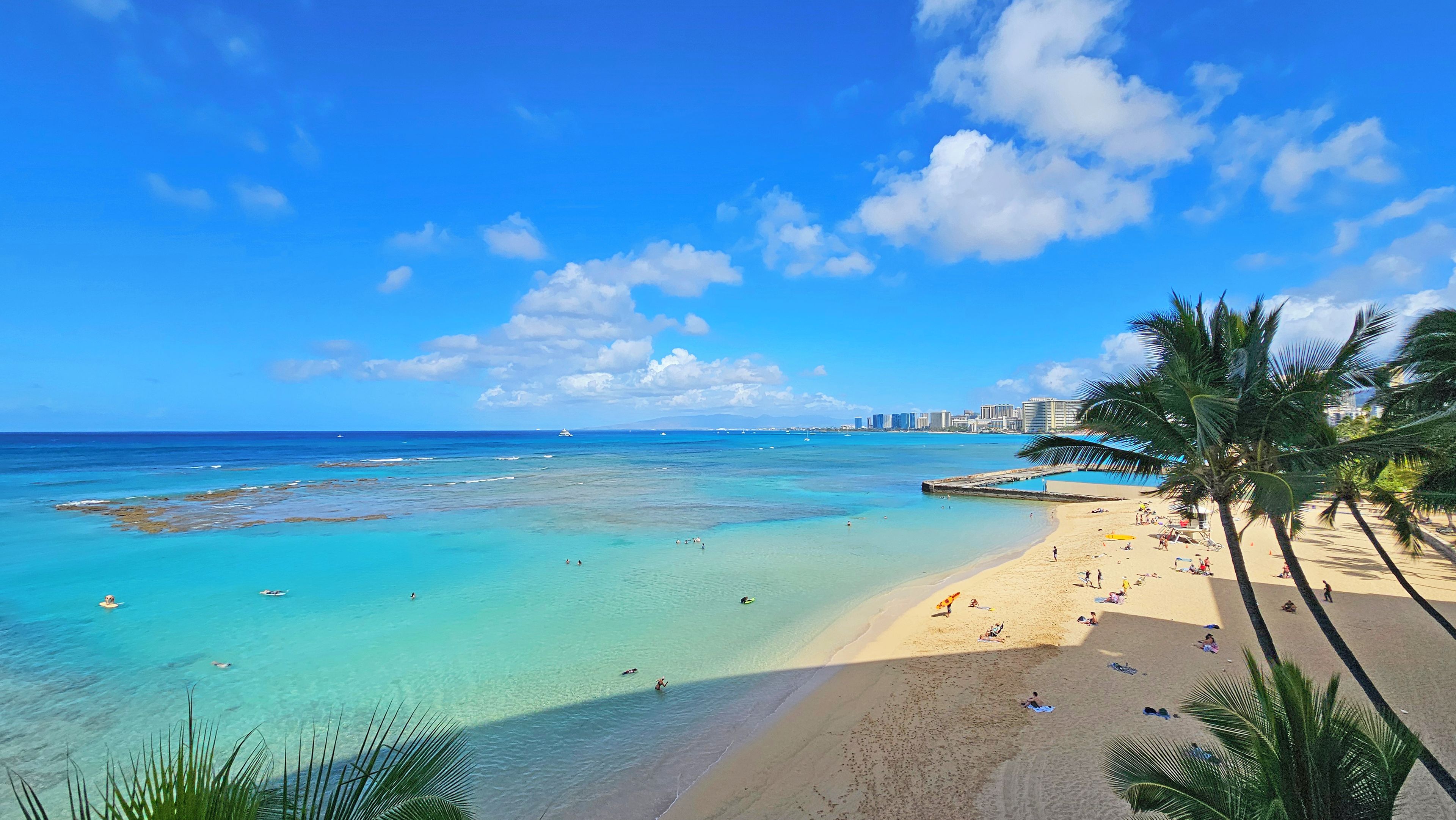 A beach with clear turquoise water, palm trees, and a distant city skyline.