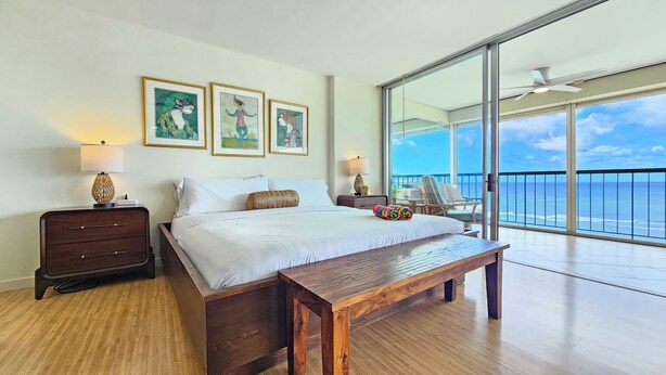 A hotel room with a large bed, a wooden bench, a nightstand, a lamp, and a view of the ocean through a balcony. - 11th Floor King Bed Beachfront Condo at Sans Souci (Honolulu)