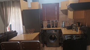 A compact kitchen with a washing machine, a coffee maker, and a stove. There are two bar stools and a dining table.