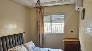 A bedroom with a bed, a nightstand, a window with blinds, a ceiling fan, and a wall-mounted air conditioner. - Modern Casablanca Apartment – Parking, A/C & WiFi (Casablanca)