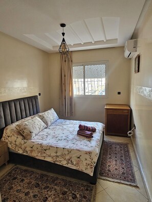 A bedroom with a bed, a nightstand, a window with blinds, a ceiling fan, and a wall-mounted air conditioner. - Modern Casablanca Apartment – Parking, A/C & WiFi (Casablanca)