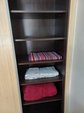 Une armoire en bois avec trois étagères ; l’étagère du haut est ornée d’un coussin coloré à rayures, l’étagère du milieu contient des vêtements pliés, et l’étagère du bas est surmontée d’une serviette rouge.
