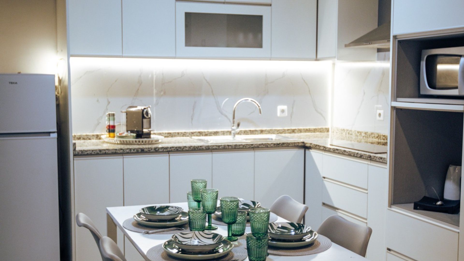 A modern kitchen with white cabinets, a microwave, a sink, and a dining table set with green glassware.