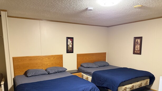 A room with two beds, each with a wooden headboard and blue bedding. There are two framed pictures on the walls. - Alliance valley Inn motel (Alliance)