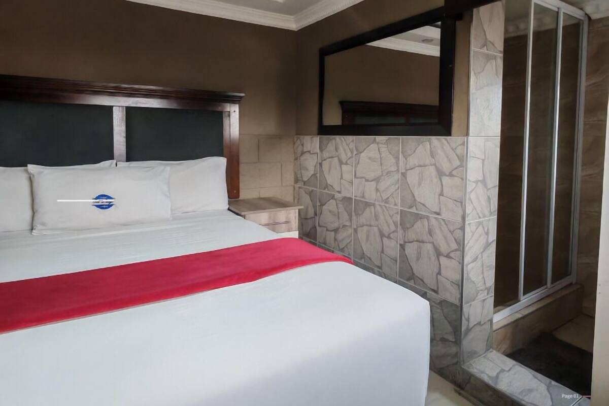 Habitación de hotel con una cama grande, una manta roja, pared de ducha con diseño de piedra y un espejo sobre la ducha.