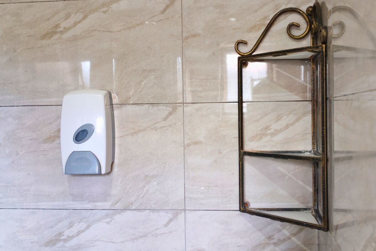 Un dispensador de gel antibacterial sujeto a una pared de azulejos junto a un marco de puerta de vidrio.