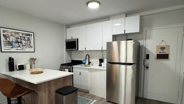 A modern kitchen with white cabinets, a stainless steel refrigerator, a microwave, and a sink. There is a wooden dining table with two chairs and a trash bin.