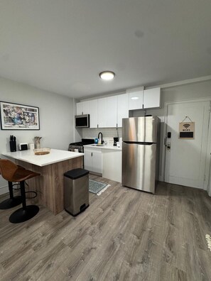 A modern kitchen with white cabinets, a stainless steel refrigerator, a microwave, and a sink. There is a wooden dining table with two chairs and a trash bin.