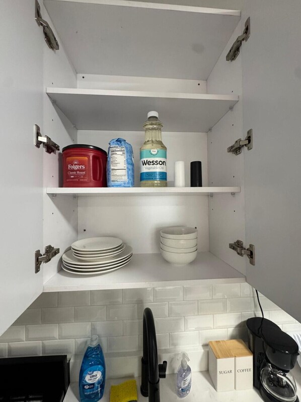 A kitchen with white cabinets, a coffee machine, sugar and coffee boxes, a dish soap bottle, a dish brush, and a Wesson vegetable oil bottle.