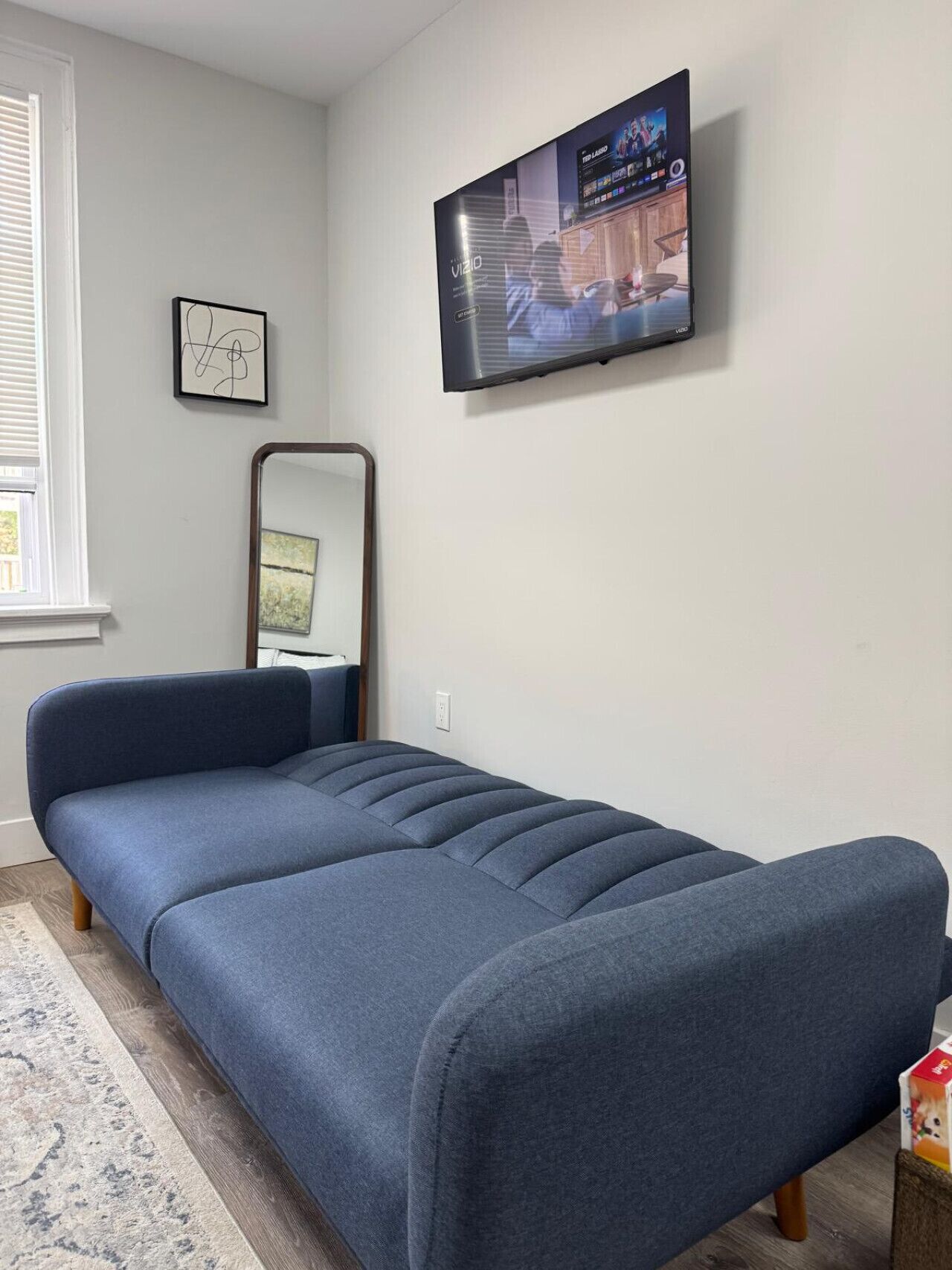 A modern bedroom with a blue sofa bed, a flat-screen TV mounted on the wall, a small framed picture, and a mirror.