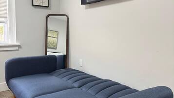 A modern bedroom with a blue sofa bed, a flat-screen TV mounted on the wall, a small framed picture, and a mirror.