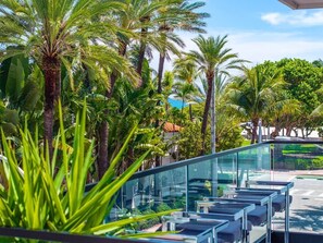 A modern outdoor area with glass railings, palm trees, and a view of the street. - Versace Mansion Neighbor (Floor 1,2,3) Sleeps 21 (Miami Beach)