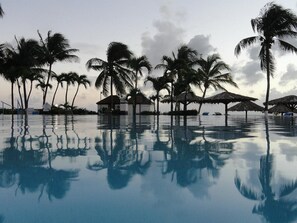 A serene pool area with palm trees and thatched-roof structures, reflecting in calm water.