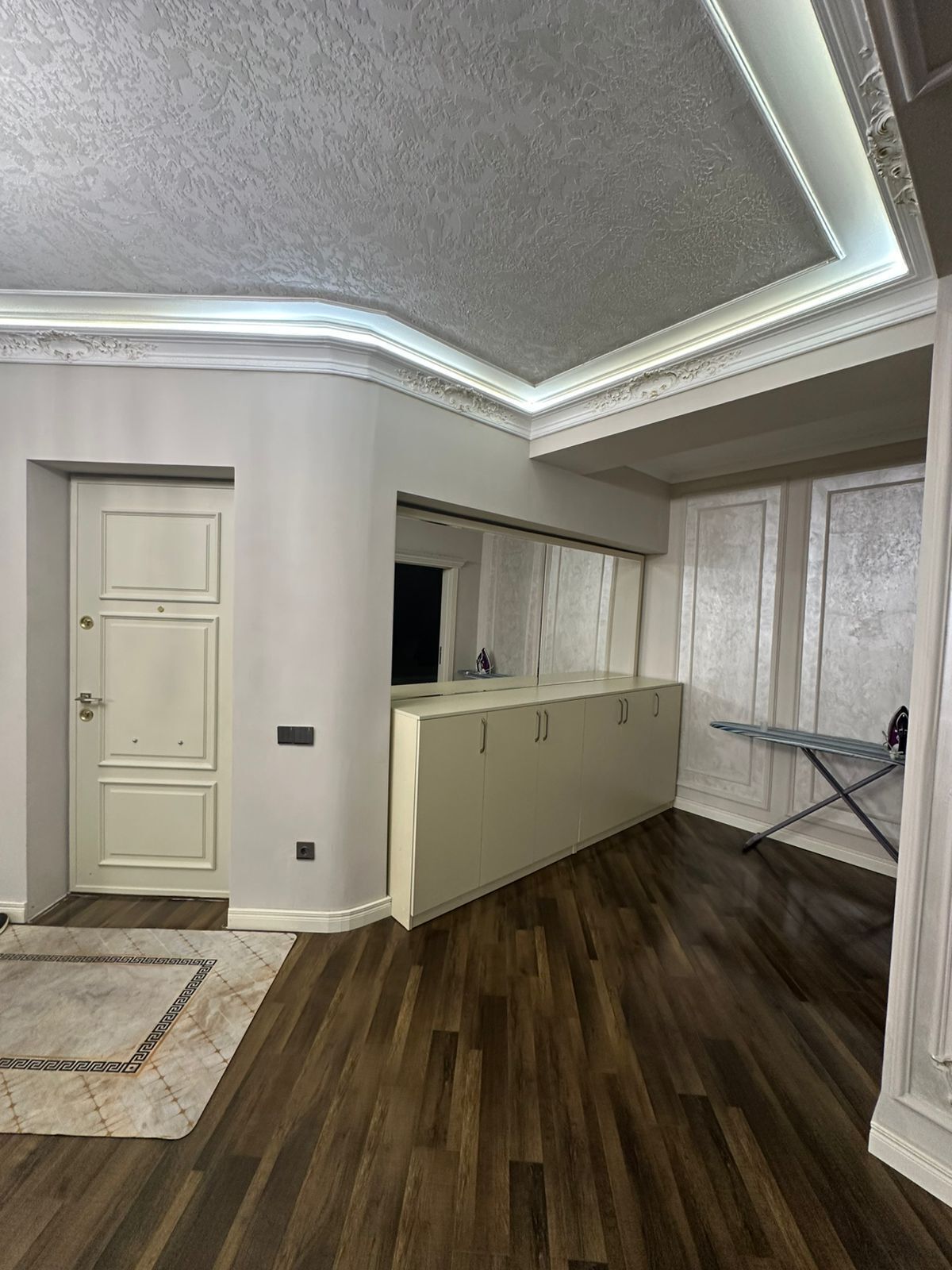 A room with a wooden floor, a white door, a white cabinet with drawers, and a small ironing board.