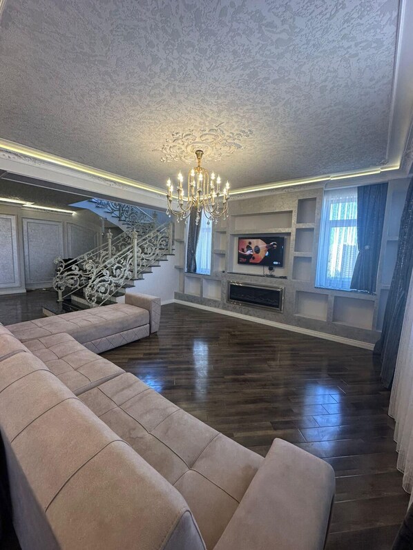Un salón amplio con un sofá seccional beige grande, una lámpara de araña, una chimenea y una escalera con barandillas ornamentales.