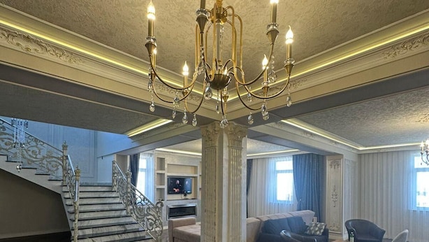 Amplia sala de estar con una imponente lámpara de araña, una escalera con barandillas ornamentadas y un mueble moderno para entretenimiento.
