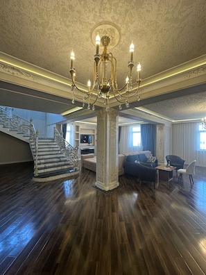 Amplia sala de estar con una imponente lámpara de araña, una escalera con barandillas ornamentadas y un mueble moderno para entretenimiento.