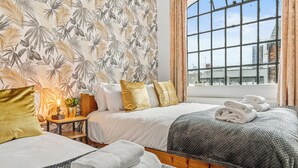 Ein Hotelzimmer mit zwei Betten, einem Fenster mit Blick auf Gebäude, einer Pflanze auf einem Nachttisch und Handtüchern auf den Betten.