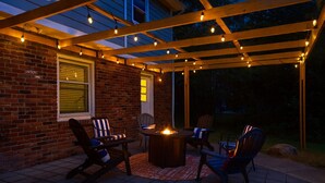 Une terrasse extérieure avec un mur en briques, une pergola en bois et des guirlandes lumineuses. On y trouve deux chaises et une table, ainsi qu’un foyer.
