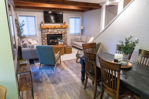 A cozy living room with a stone fireplace, a sofa, a dining table with chairs, and a staircase.