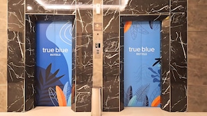 Kaksi hissin ovea, joissa on näkyvillä hotelliketju True Blue Hotelsin logo, marmoriseinät ja keskeinen hissin ohjauspaneeli.