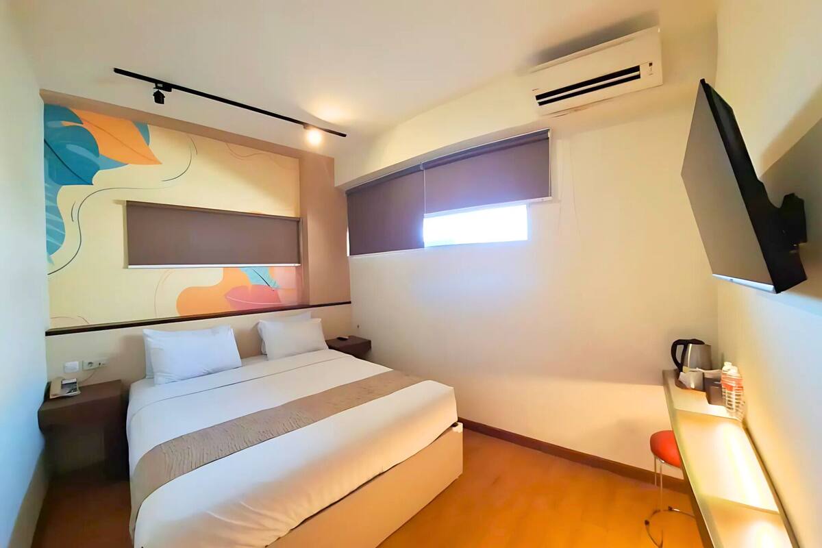 A hotel room with a bed, bedside table, air conditioning unit, window, and a TV mounted on the wall.