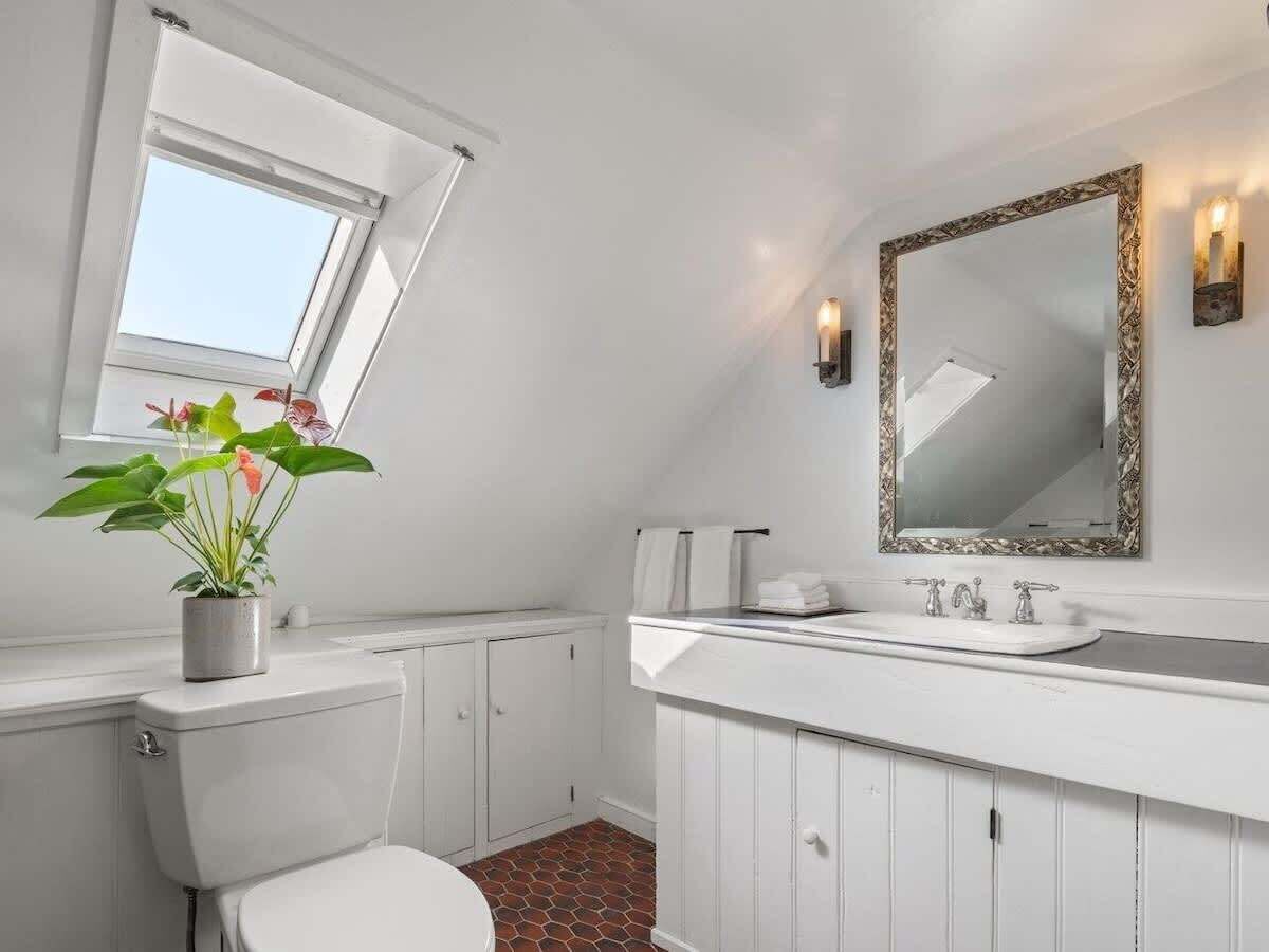 Une salle de bain avec des toilettes blanches, un lavabo avec un miroir, une plante en pot et une fenêtre.