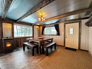 Un intérieur rustique avec une table et des bancs en bois, une cheminée en pierre et de grandes fenêtres donnant sur la forêt.