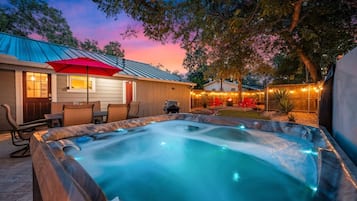 A hot tub with a wooden deck and a patio area with chairs and a red umbrella.