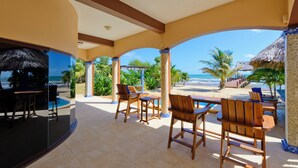 Un espacio exterior techado con mobiliario de madera, una piscina y vistas a la playa y al océano.