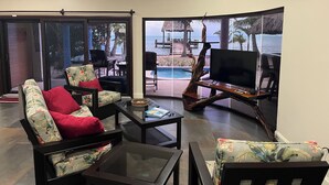 Sala de estar con sofá, mesa de centro y televisor, con vista a una piscina y zona de playa.