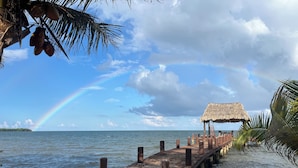 Un muelle de madera que se adentra en aguas tranquilas, una estructura con techo de paja y un arcoíris visible.