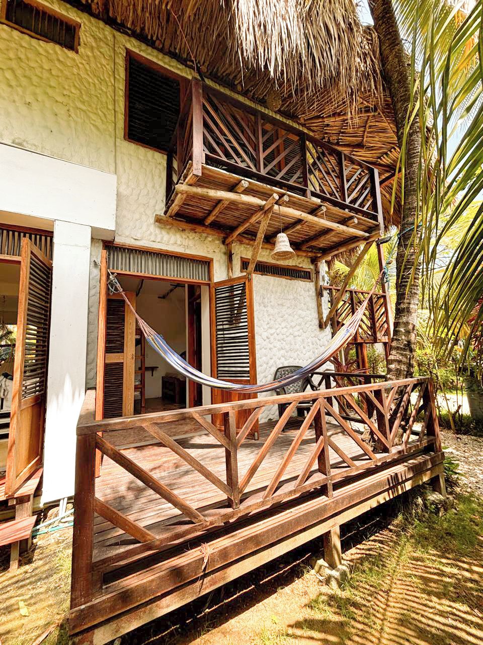 A wooden deck with a hammock, a thatched roof, and a white building with wooden shutters.
